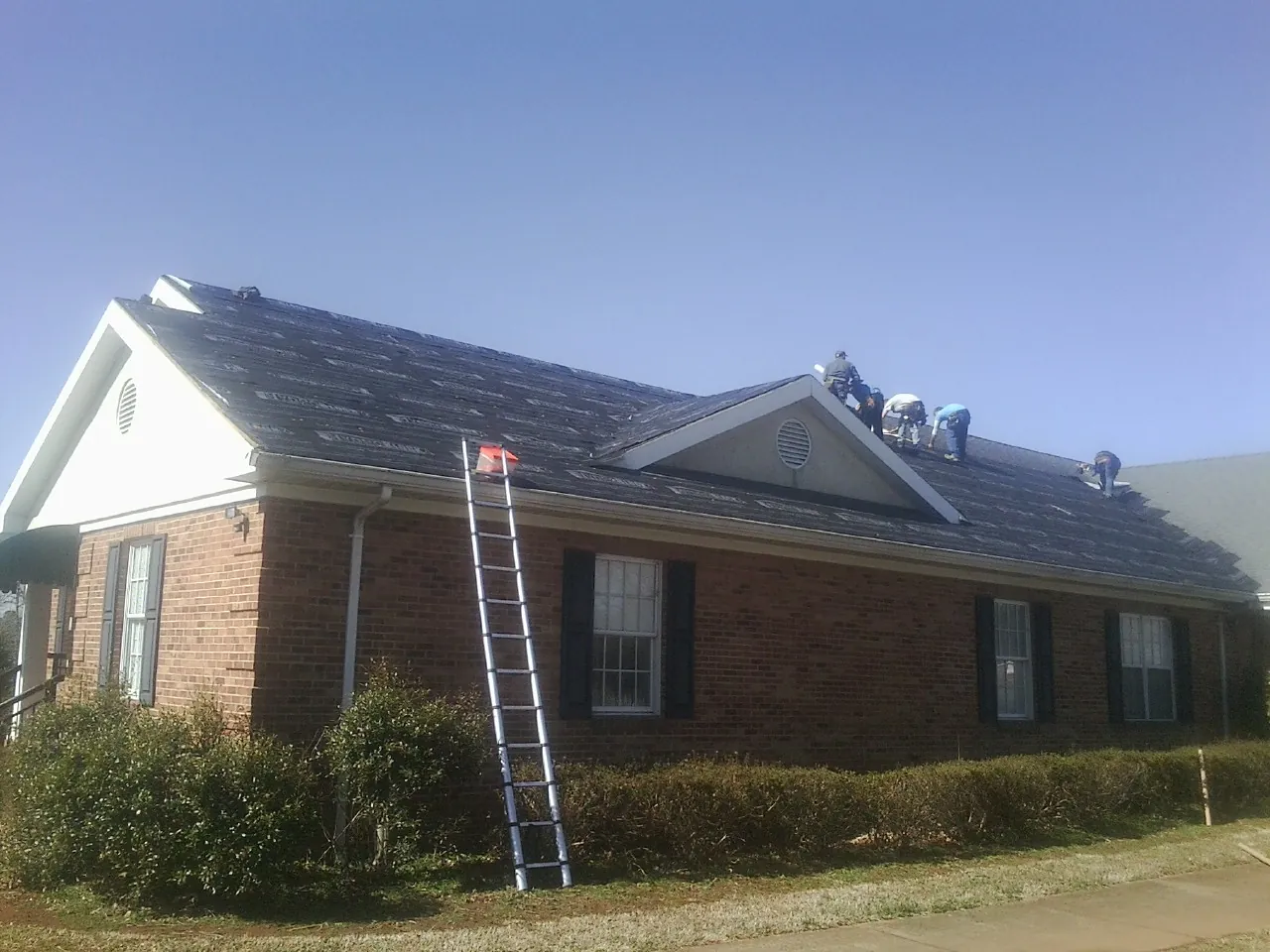 Crew tearing shingles off a brick ranch roof with a ladder set — Emergency Roof Repair work by our crew in Brave