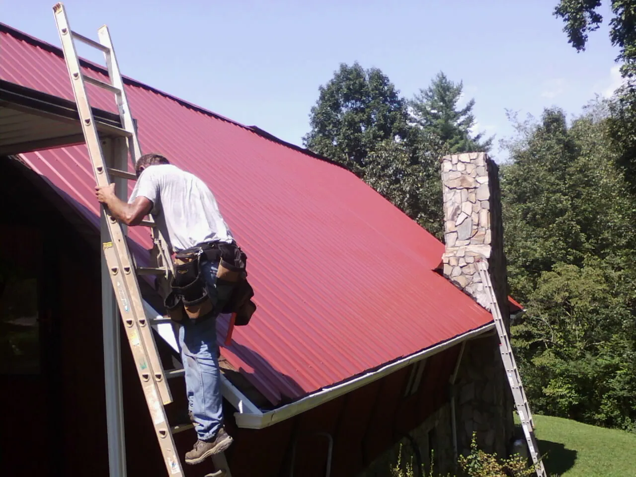 Roofer on a ladder beside a red metal roof and stone chimney — Soffit & Fascia Repair by Roofing Brothers in Brave